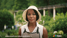 a woman wearing a straw hat and apron is standing in a garden with a nbc logo in the corner