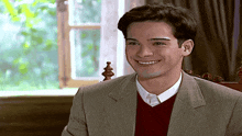 a man in a suit and red sweater smiles in front of a window .