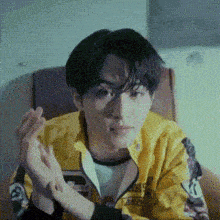 a young man in a yellow jacket is sitting in a chair with his hands together .