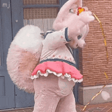 a stuffed animal in a pink dress is standing in front of a brick building holding a ribbon .