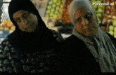 two women wearing hijabs are standing next to each other in front of fruits .