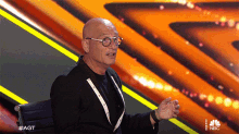 a bald man wearing glasses and a black jacket is sitting in front of a nbc logo