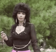a woman in a black top and pink skirt is standing in a park holding a cigarette .