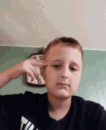 a young boy is pointing his finger at his forehead in front of a clock that says 11 12