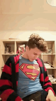 a man wearing a superman t-shirt and a red and black striped jacket is sitting in front of a bookshelf .