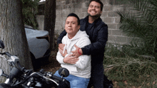 a man on a motorcycle is hugging another man in a white hoodie