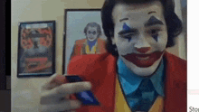 a man in a clown costume is holding a cell phone in front of a picture of the joker .