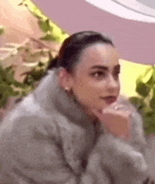 a woman wearing a fur coat and earrings is sitting down with her hand on her chin .