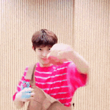 a young boy in a pink striped shirt is standing in front of a wooden wall holding a bag .