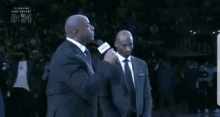 a man in a suit is talking into a microphone while another man stands behind him with the number 8 on the screen
