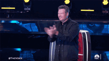 a man in a black shirt applauds while sitting in a chair with the words #thevoice on the bottom