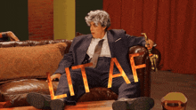 a man in a suit and tie is sitting on a couch with lit af written on his pants