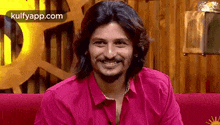 the man is wearing a pink shirt and smiling while sitting on a red couch .