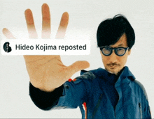 a man wearing glasses and a blue jacket holds up his hand in front of a hideo kojima repost