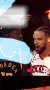 a man in a rockets jersey is standing next to another man in a black hat .