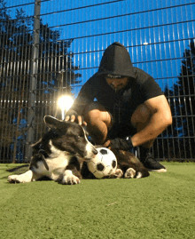 a man in a black hoodie kneeling next to a dog with a soccer ball in its mouth