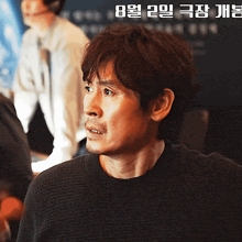 a man in a black sweater is looking to the side with a foreign language advertisement behind him
