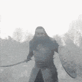 a man with long hair is holding a sword and shield in a foggy field .