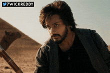 a man with a beard is standing in the desert with a camel in the background and the words 7wickreddy below him