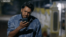 a man in a denim shirt is looking at his phone with xtomsj2005 written on the bottom right