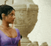 a woman in a purple top and necklace stands in front of a statue