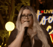 a woman wearing glasses is singing into a microphone in front of a live sign .