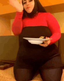 a woman in a red and black dress is kneeling down holding a plate of food .