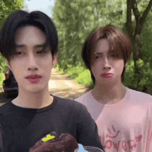 two young men are standing next to each other in a park . one of the men is holding a piece of chocolate cake .