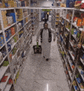 a man in a white robe is walking down a aisle in a grocery store holding a green basket