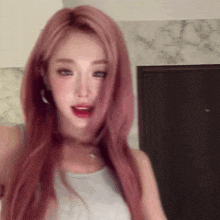 a woman with long pink hair is wearing a white tank top and earrings and looking at the camera .