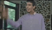 a man in a purple shirt is standing in front of a window with his hand outstretched .