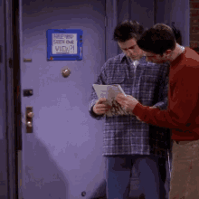 two men are standing next to each other in front of a purple door looking at a magazine .