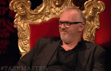 a man in a suit and glasses is sitting in a chair with the hashtag taskmaster