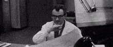 a black and white photo of a man in a lab coat sitting at a desk looking at a drawing .