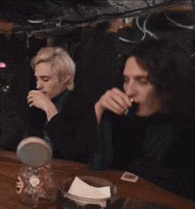 a man drinking from a shot glass next to another man with a sign on the wall that says mixing room