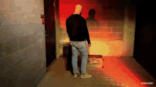 a bald man in a black sweater stands in a hallway with a red light behind him