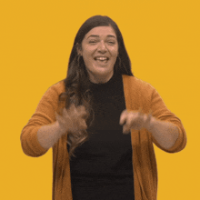 a woman wearing a black shirt and an orange cardigan is smiling and waving