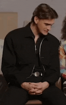 a man in a black jacket is sitting down with his hands folded .