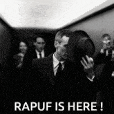 a black and white photo of a man in a suit holding his hat with the caption " rapuf is here "