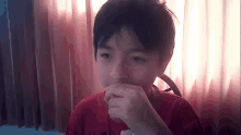 a young boy in a red shirt is sitting in front of a window and covering his mouth with his hand .
