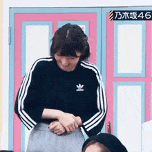 a woman wearing a black adidas jacket is standing in front of a pink door and smiling .