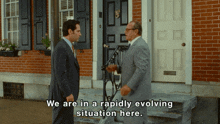 two men in suits are standing in front of a brick building and one of them says we are in a rapidly evolving situation here