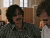 a man with curly hair and a mustache is wearing a plaid shirt with the letter t on the front