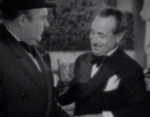 a man in a tuxedo talks to another man in a hat in a black and white photo