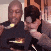a man is eating a bowl of food with a fork and spoon while another man is eating a bowl of food .
