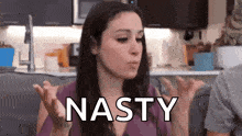 a woman is sitting on a couch in a kitchen with her hands up and the word nasty written on her face .