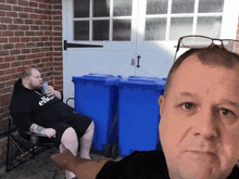 a man wearing a black ellesse shirt sits in front of two blue trash cans
