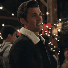 a man in a black jacket with a white fur collar is standing in front of a string of lights .