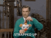 a man in a green sweater is holding a stuffed animal with the words `` strapped up '' written below him .