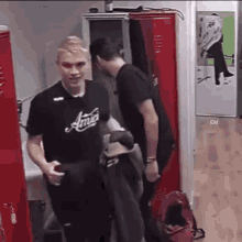 two men are standing next to each other in a locker room . one of the men is wearing a black shirt that says amici